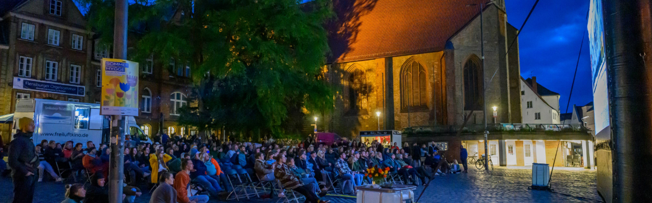 Menschen sitzen abends auf Klappstühlen in einem historischen Innenhof und schauen einen Film auf einer großen Open‑Air‑Leinwand, umgeben von warm beleuchteten Backsteingebäuden.