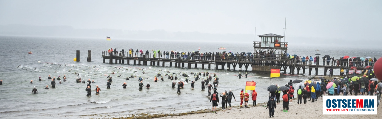 Zahlreiche Triathleten in schwarzen Neoprenanzügen starten beim Schwimmen im grauen Ostseewasser. Rechts im Bild ist eine Seebrücke voller Zuschauer mit Regenschirmen zu sehen. Unten rechts befindet sich das Logo des 'OstseeMan Triathlon Glücksburg'.