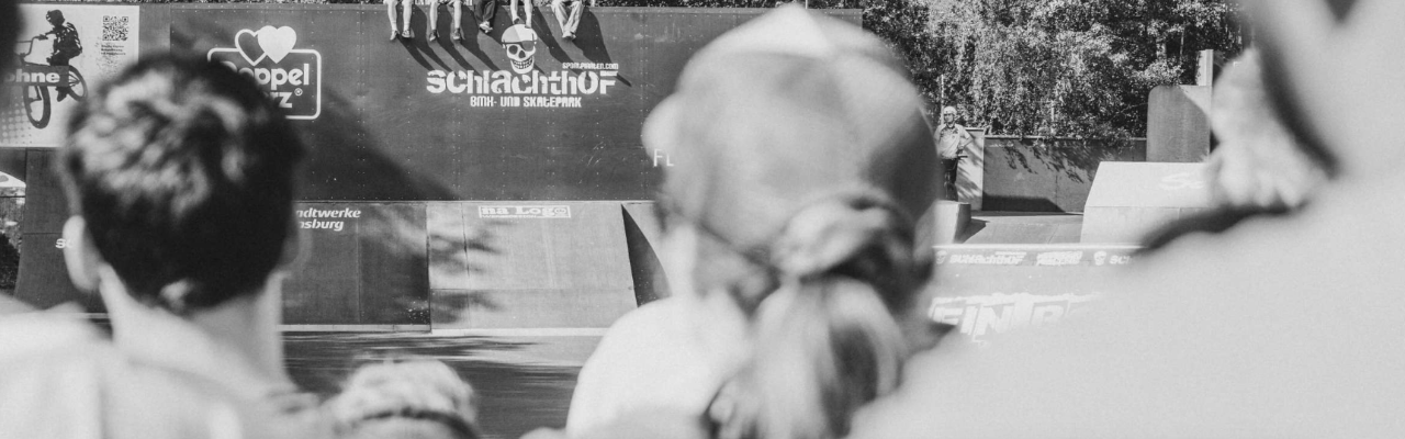 Zuschauer stehen vor einer BMX‑ und Skatepark‑Rampenanlage mit großem Schlachthof‑Schriftzug, während im Hintergrund ein Outdoor‑Event stattfindet. Das Foto hat einen schwarz-weiß Filter.