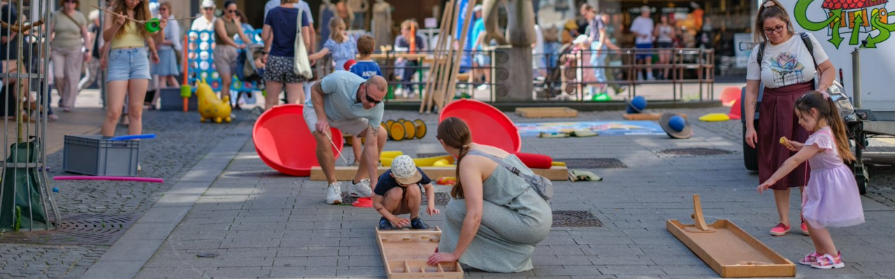 Kinder und Erwachsene spielen auf einer Fußgängerzone an mehreren Holz‑Geschicklichkeitsspielen, während im Hintergrund weitere bunte Outdoor‑Spielstationen eines Familienfestes aufgebaut sind.