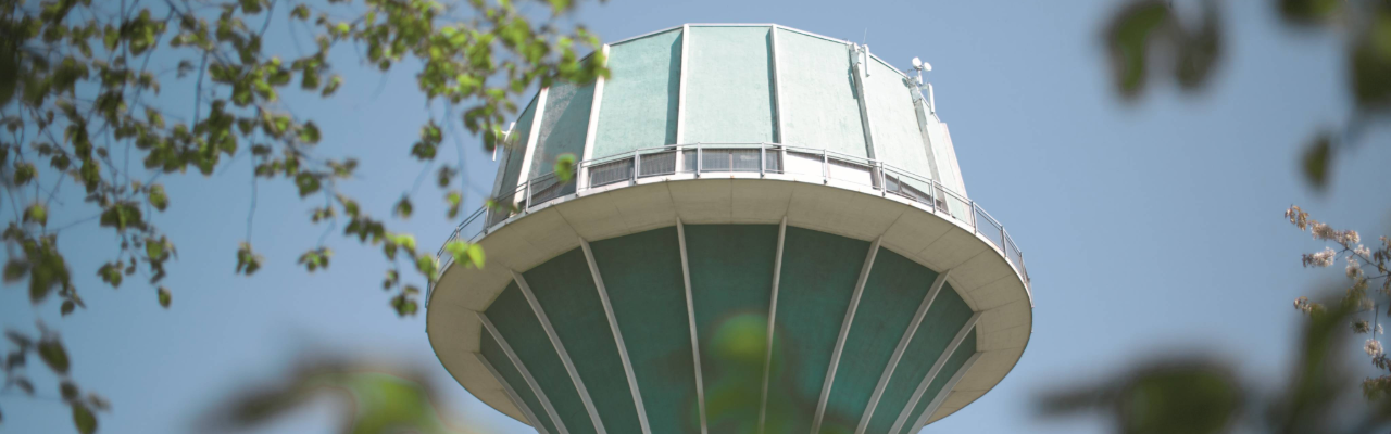 Oberer Bereich eines Wasserturms mit Aussichtsplattform vor blauem Himmel und einigen Ästen am Rand des Bildes.