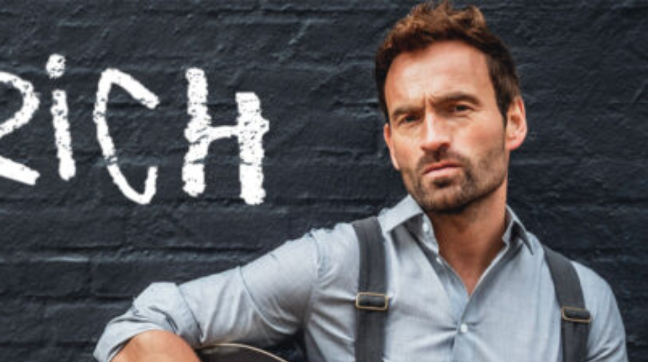 Junger Mann mit braunen Haaren und Bart mit blauen Hemd und Hosenträgern steht vor einer schwarzen Mauer mit der weißen Schrift rich.