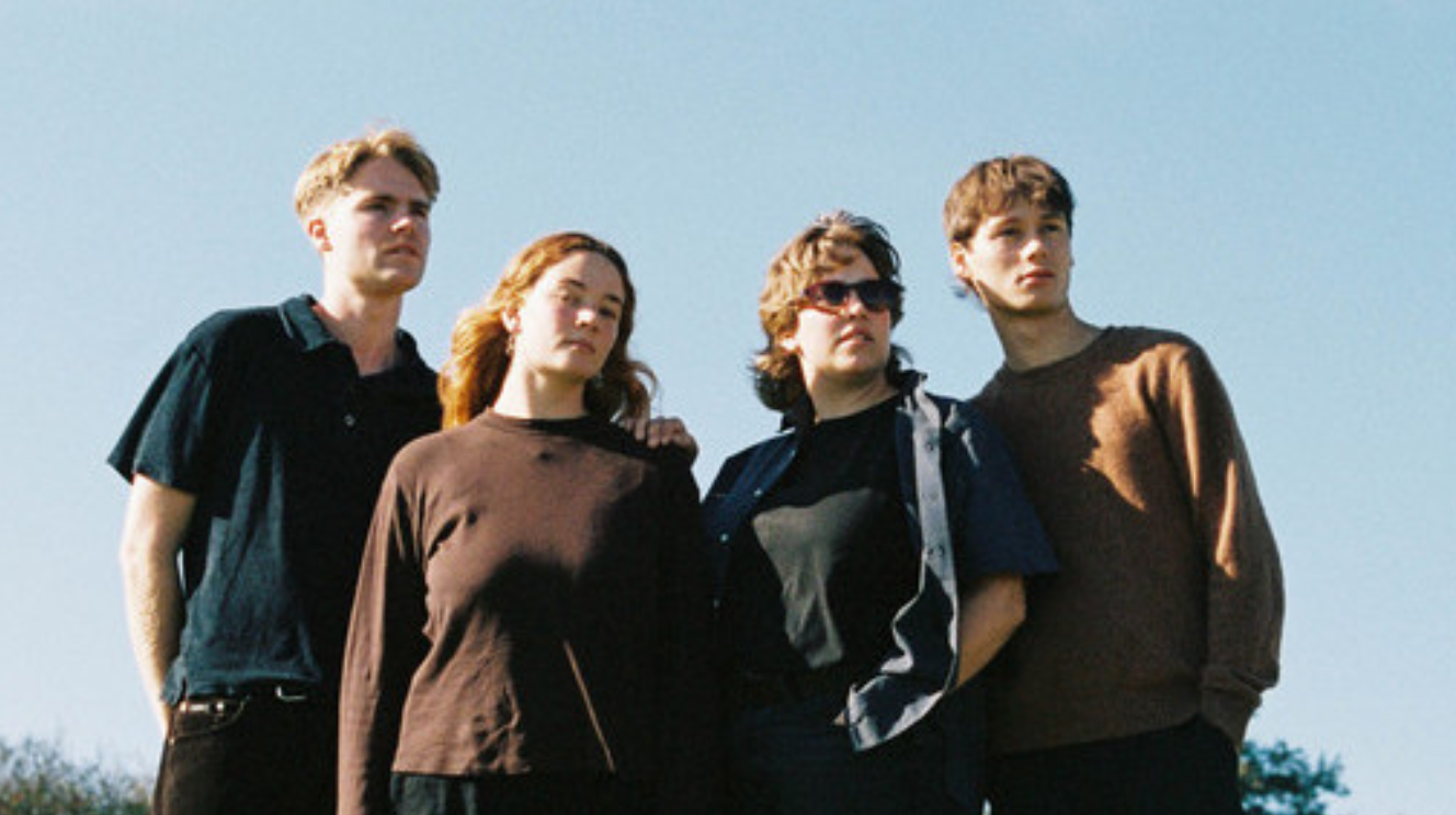 3 junge Männer und eine junge Frau stehen vor blauen Himmel und schauen von schräg oben Richtung Kamera. Sie tragen dunkle, eher lockere Kleidung. 
