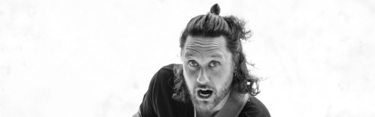 Mann mit kinnlagen Haaren und einem halb zurückgebundenen Dutt schaut mit offenem Mund in die Kamera. Er trägt ein schwarzes Shirt und markanten Bart.