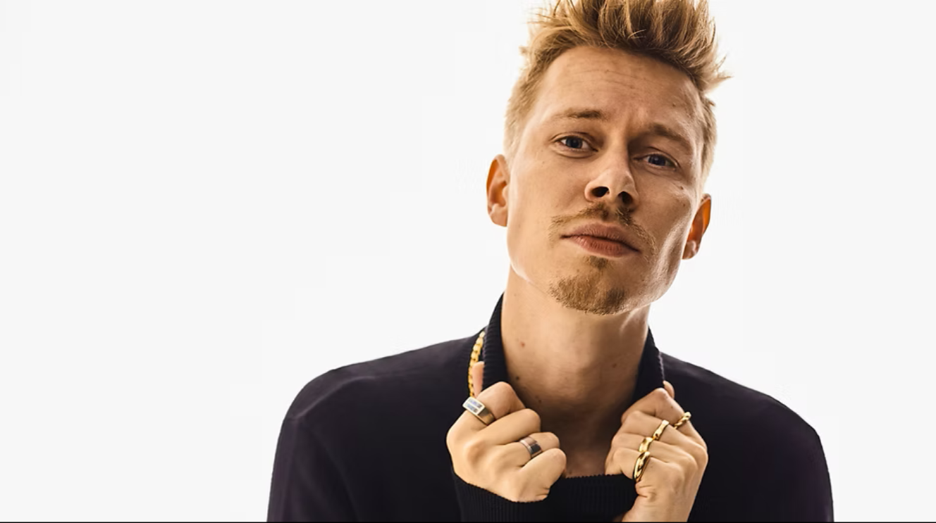 Blonder junger Mann vor weißem Hintergrund schaut mit ernster Mine in die Kamera. Dabei hält er die Hände am Kragen seines schwarzen Oberteils. Er trägt bis auf an zwei Fingern Ringe.