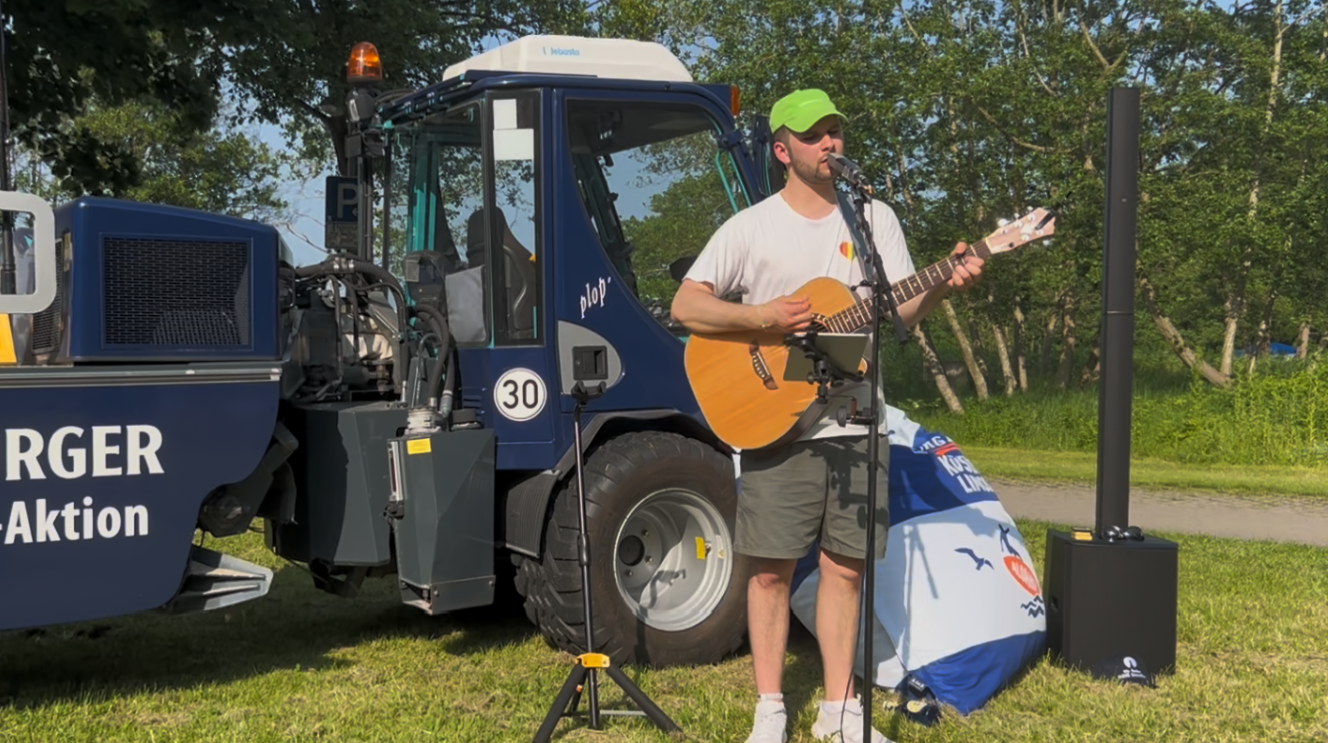 Junger Mann mit grüner Cap, weißem Shirt und kurzer Shorts spielt auf einer Gitarre. Im Hintergrund sind Bäume und Rasen zu sehen sowie ein blauer Kleintraktor mit der Aufschrift plop, 30 und RGER Aktion.