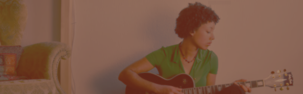 Seitliches Profil einer jungen Dame mit dunklen Haaren, die auf einer Gitarre spielt. Links im Bild ist ein Teil eines alten Sofas zu sehen.Das Foto hat einen starken Filter, der das Bild verbäunlicht.