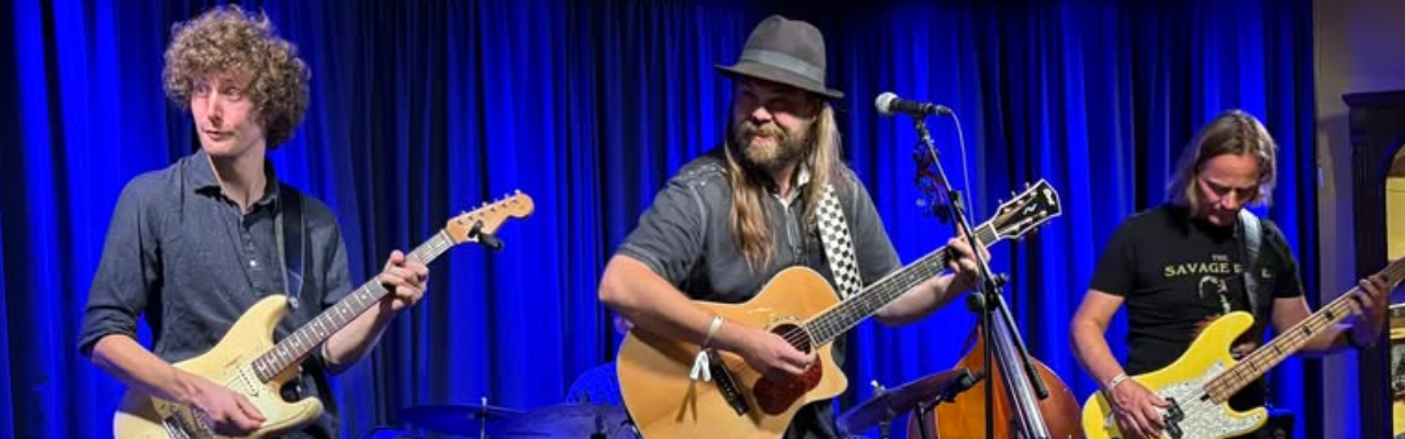 Drei Musiker spielen auf einer Bühne vor blauem Vorhang: links ein Gitarrist mit lockigen Haaren an einer E-Gitarre, in der Mitte ein Sänger mit Hut an einer Akustikgitarre und Mikrofon, rechts ein Bassist mit E-Bass.