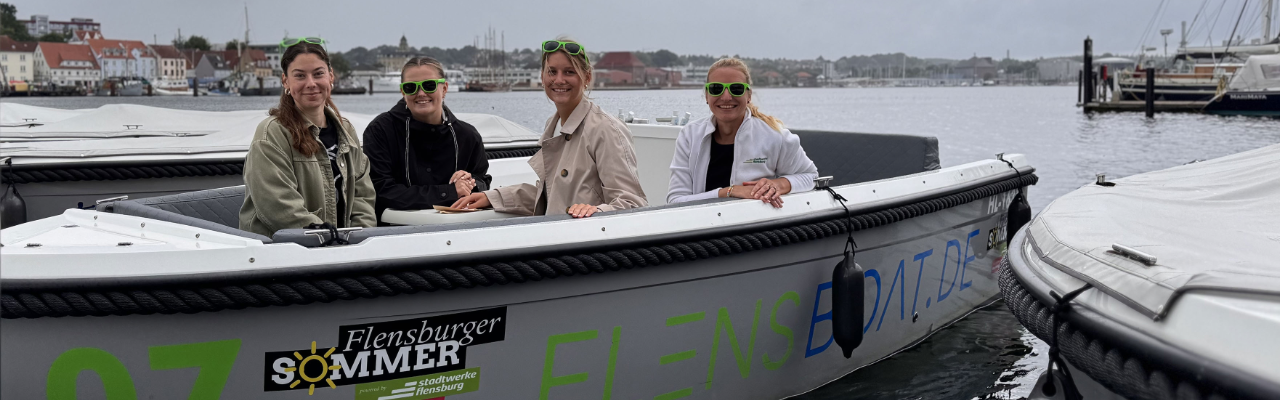 Graues Boot auf dem Wasser mit Aufklebern Flensburger Sommer, Stadtwerke Flensburg und FlensBoat. 4 freundlich schauende junge Frauen sitzen mit grüner Sonnenbrille im Boot.