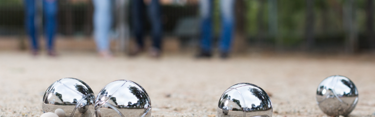 Mehrere Metall- und Holzkugeln liegen auf einem sandigen Spielfeld, während im Hintergrund unscharf Personen stehen.