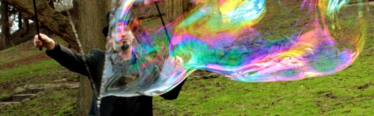 Mann im Freien, die mit zwei Stäben und einer Schnur eine riesige, farbig schillernde Seifenblase auf einer Wiese zwischen Bäumen erzeugt.