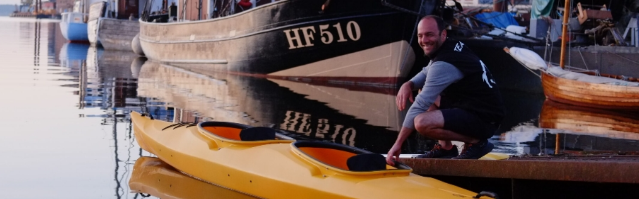 Mann hockt neben einem gelben Doppelkajak an einem ruhigen Hafen, umgeben von mehreren festgemachten Booten.