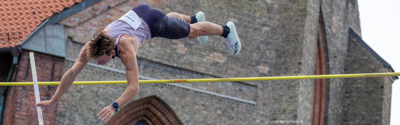 Muskulöser Stabhochspringer in der Luft beim Überqueren der Latte während eines Wettkampfs vor einem Backsteingebäude.