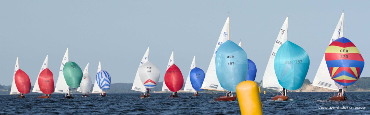 Mehrere Segelboote mit farbigen Spinnakern segeln während einer Regatta über ruhiges Wasser, im Vordergrund eine gelbe Boje.