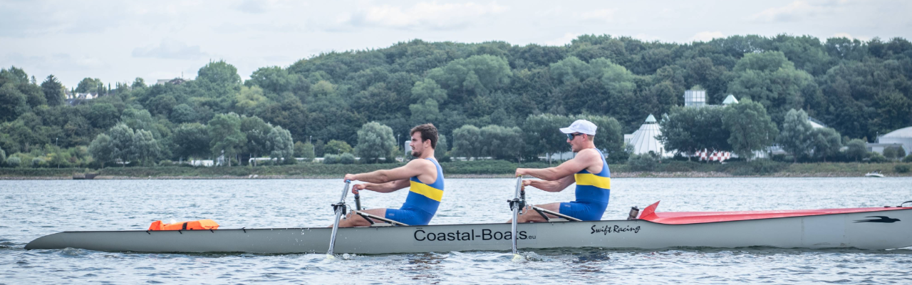 Zwei Ruderer in blau‑gelben Wettkampfanzügen bewegen ein schmales Rennboot synchron über ruhiges Wasser, vor einer bewaldeten Uferkulisse. Auf dem Boot steht die Aufschrift Coastal-Boats.