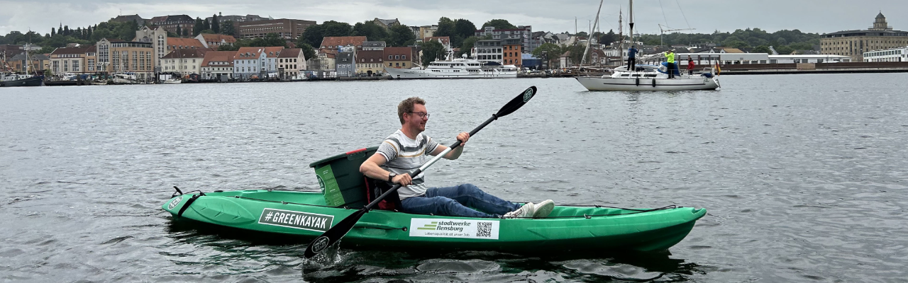 Greenkayak mit Stadtwerke Flensburg Logo und einer großen Tonne in der Mitte auf dem Wasser. Ein junger Mann sitzt in der Mitte und hält ein Paddel in der Hand.