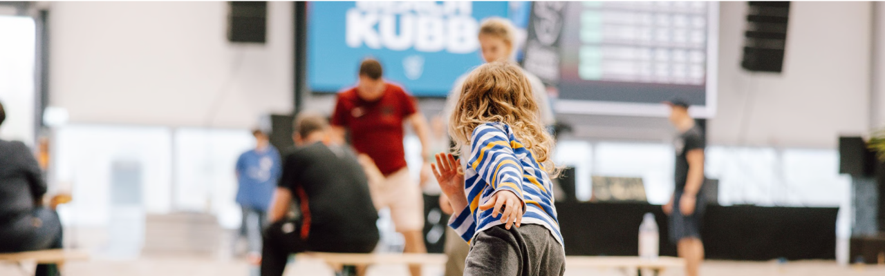 Kind wirft in einer Halle ein Spielholz, während Erwachsene im Hintergrund an einem Kubb‑Spiel teilnehmen.