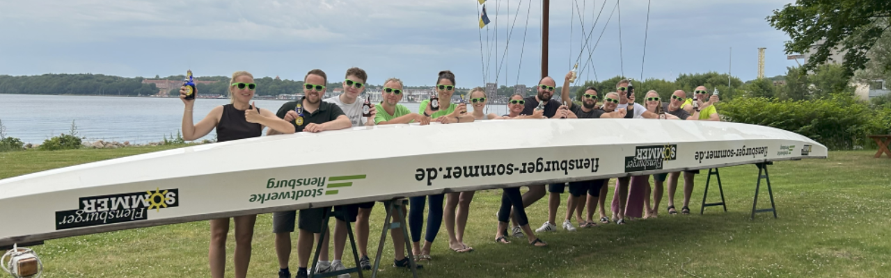 Gruppe von Menschen hebt im Freien ein großes weißes Ruderboot an, alle tragen grüne Sonnenbrillen, im Hintergrund Wasser, Bäume und Gebäude.
