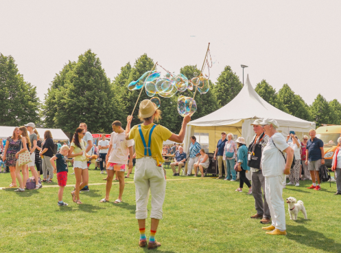 Straßenkünstler auf einer Wiese, der mit zwei Stäben große Seifenblasen erzeugt, umgeben von zuschauenden Kindern und Erwachsenen auf einem belebten Outdoor‑Fest mit Zelten im Hintergrund.