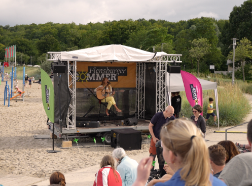 Open‑Air‑Bühne am Strand mit einem Musiker, der akustische Gitarre spielt, während Besucher auf Bänken sitzen und das Sommer‑Event Flensburger Sommer verfolgen.