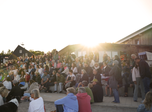 Große Menschenmenge sitzt draußen auf Stufen und blickt auf eine Bühne oder Veranstaltung, während die tief stehende Sonne hinter Gebäuden einen warmen Lichtschein über die Szene legt.