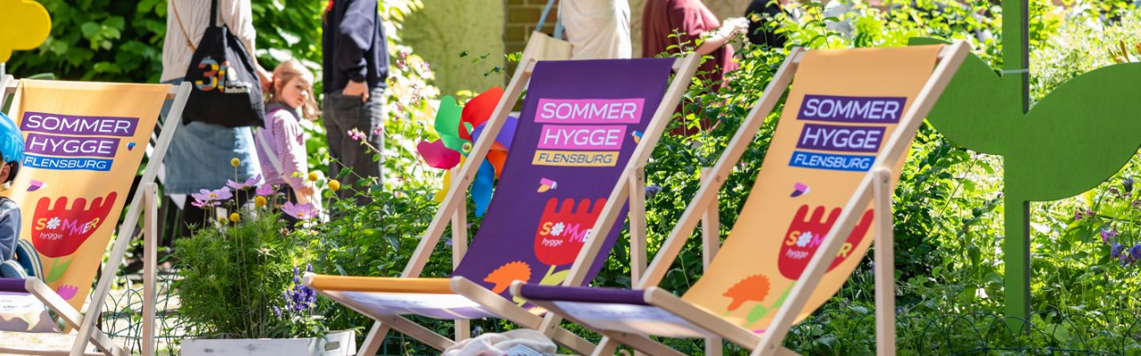 3 bunte Liegestühle mit dem Aufdruck Sommer Hygge stehen vor blühenden Blumen. Im Hintergrund ist eine Schlange an Menschen zu erkennen.