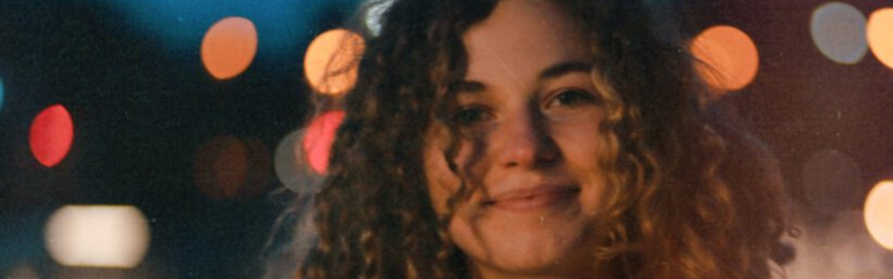 Ausschnitt eines Porträts eines lächelnden Mädchens mit braunen Locken. Der Hintergrund ist schwarz mir bunten Kreisen.