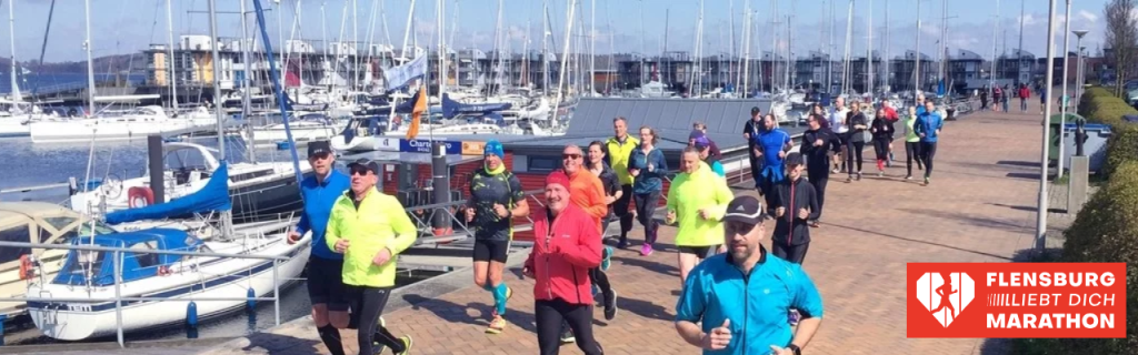Eine Gruppe von Läufern in bunter Sportkleidung joggt bei sonnigem Wetter an der Hafenpromenade entlang. Im Hintergrund liegen zahlreiche Segelyachten an den Stegen. Unten rechts befindet sich das rote Logo des 'Flensburg liebt dich Marathon'.